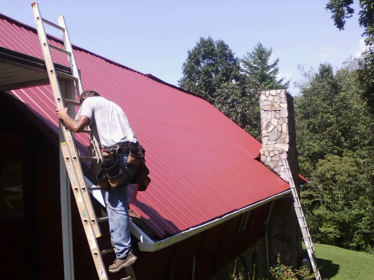 Roofer on a ladder beside a red metal roof and stone chimney — Soffit & Fascia Repair by Roofing Brothers in Weaverville